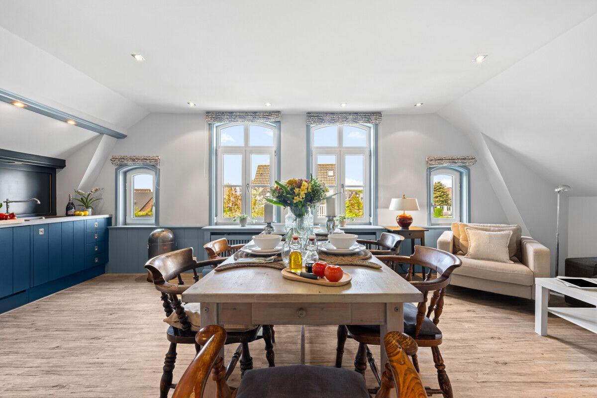 Helles Wohn-Esszimmer mit Holztisch, blauen Küchenzeilen und Sofa unter drei Fenstern.