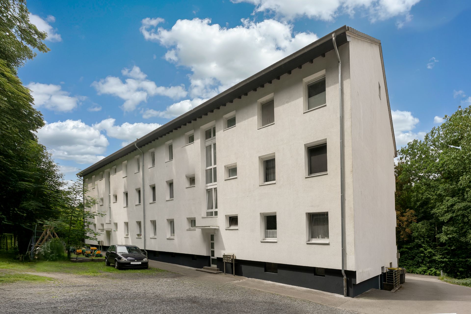 Weißes Mehrfamilienhaus mit mehreren Fenstern, Auto parkt davor, Bäume im Hintergrund.