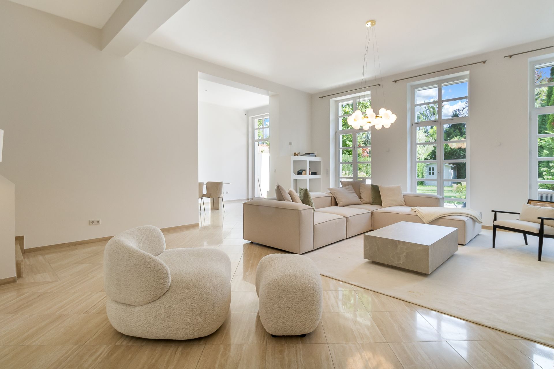 Helles Wohnzimmer mit Ecksofa, zwei Sesseln, Couchtisch und großen Fenstern mit Blick nach draußen.