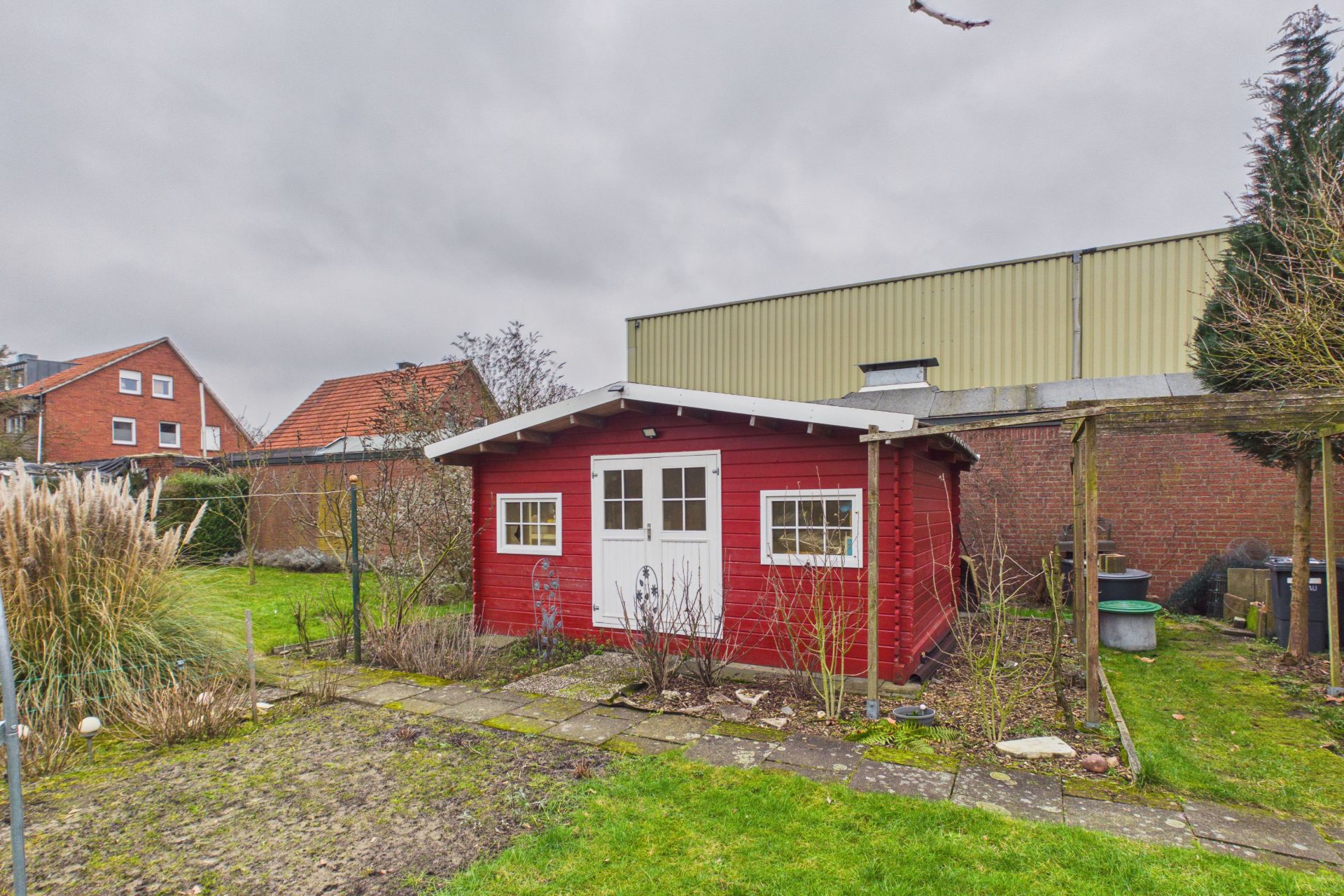 Rotes Gartenhaus mit weißen Türen und Fenstern steht in einem grünen Garten.