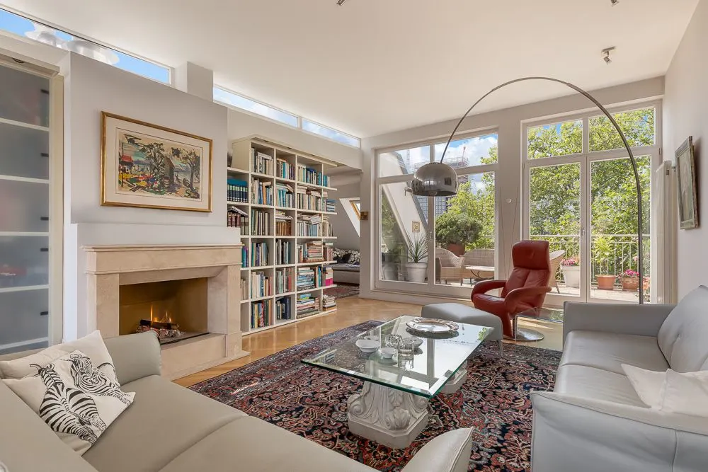 Helles Wohnzimmer mit Sofa, Kamin, Bücherregal, Glastisch und Zugang zum Balkon.