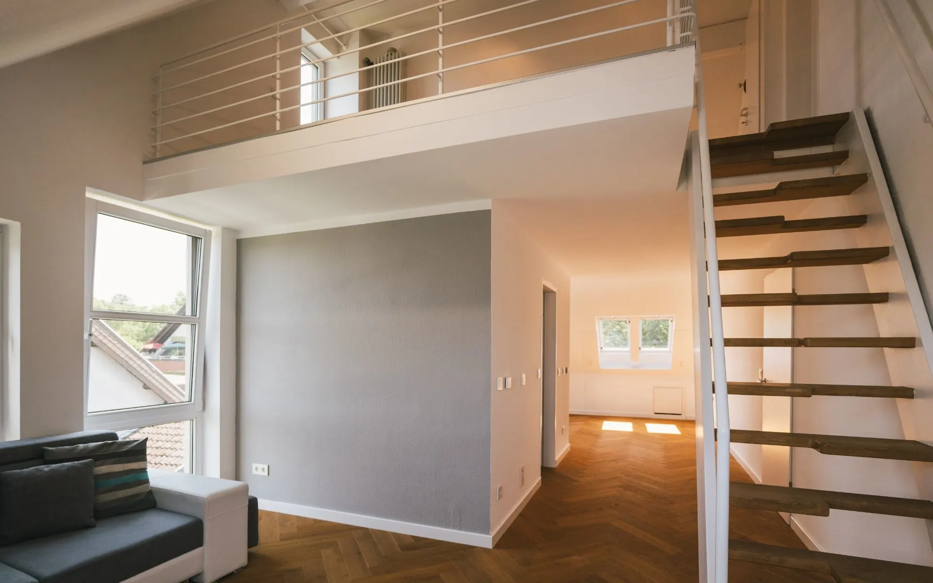 Wohnzimmer mit Couch, Fenster, Holztreppe und offener Galerie auf der oberen Etage.