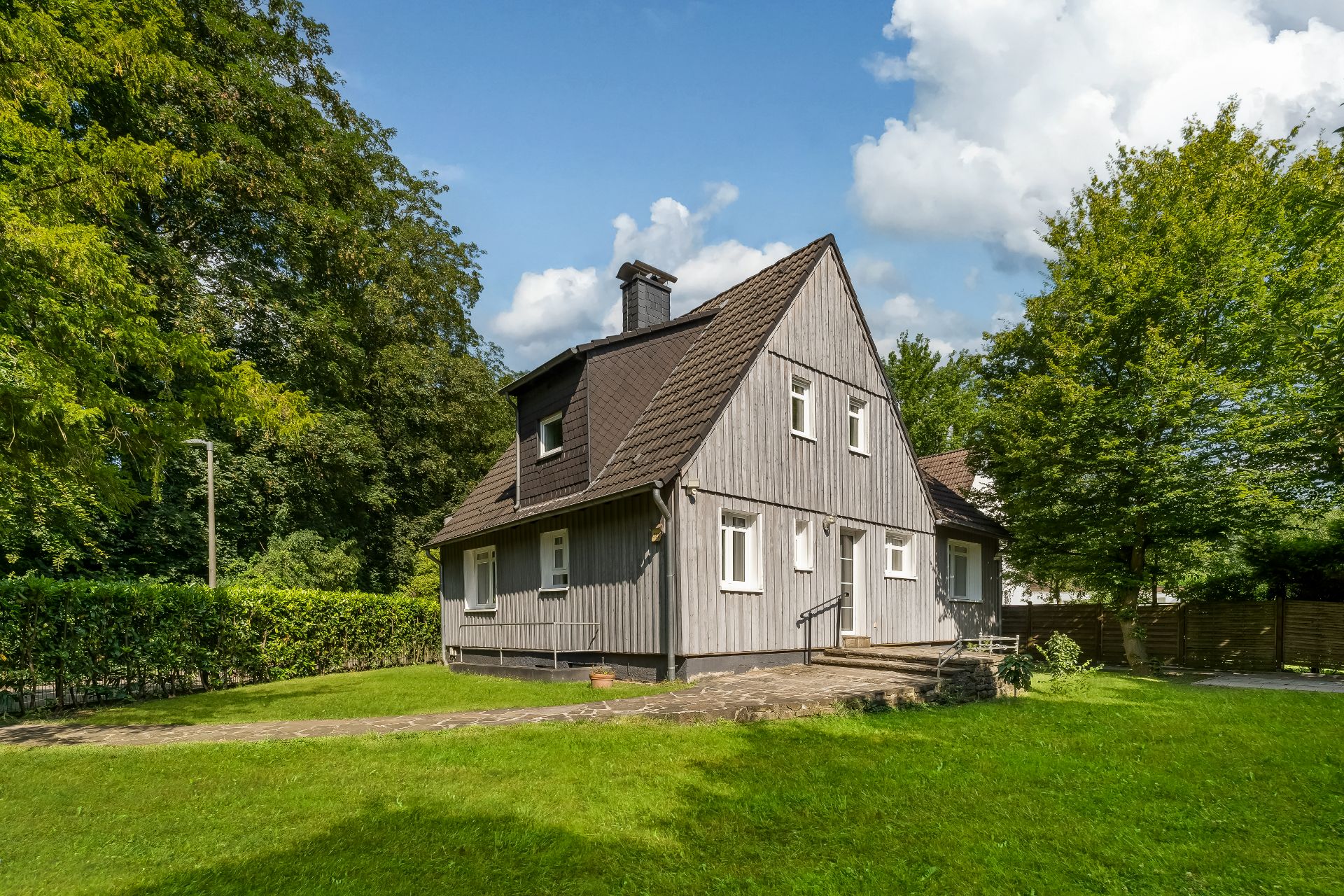 Graues Holzhaus mit Satteldach und kleinem Garten, umgeben von Bäumen.