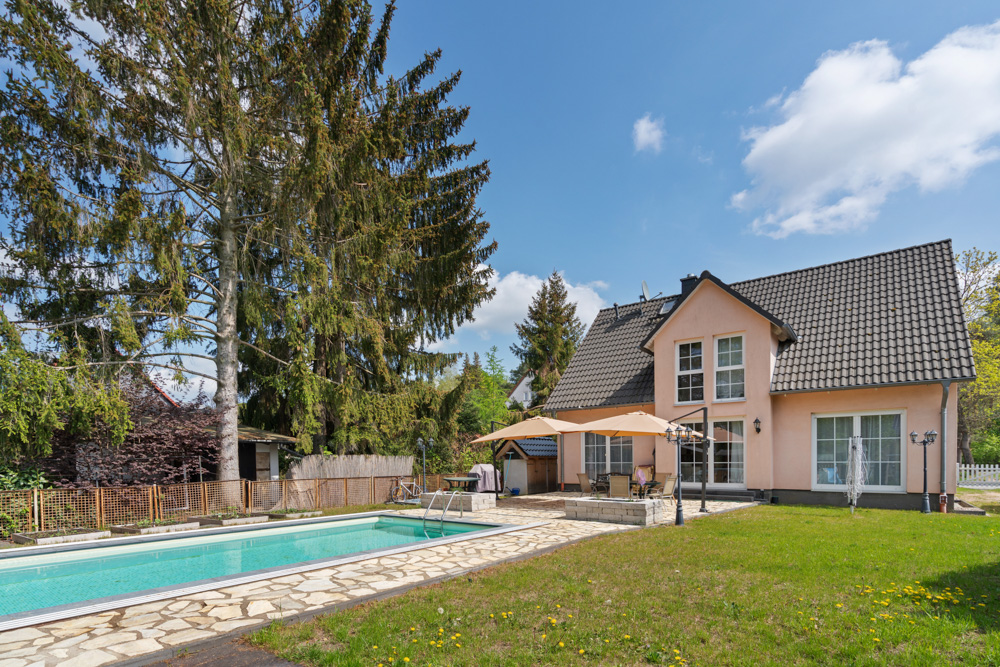 Haus mit Garten, Terrasse, Swimmingpool und großen Bäumen im Hintergrund bei sonnigem Wetter.
