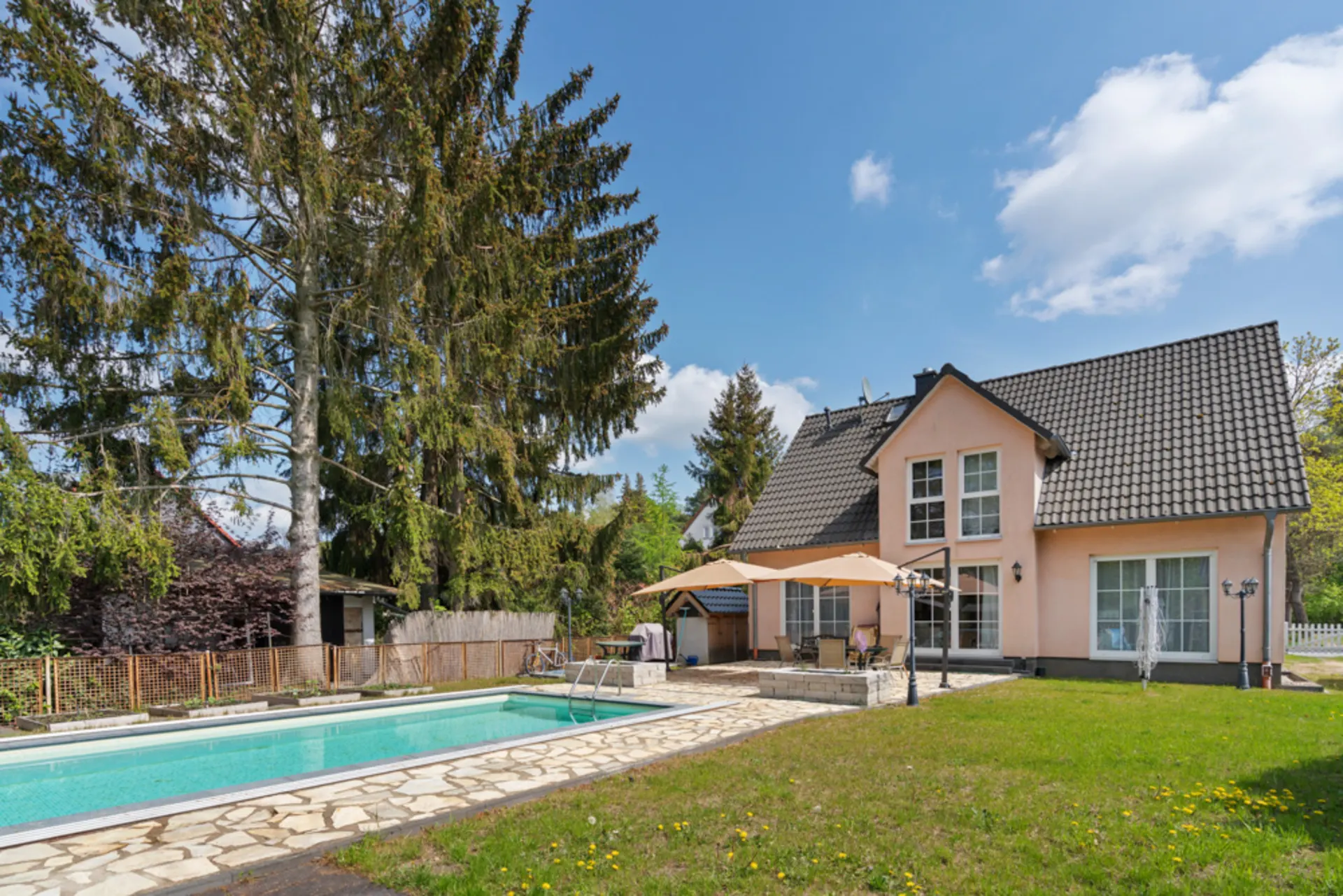 Haus mit Garten, Terrasse, Swimmingpool und großen Bäumen im Hintergrund bei sonnigem Wetter.