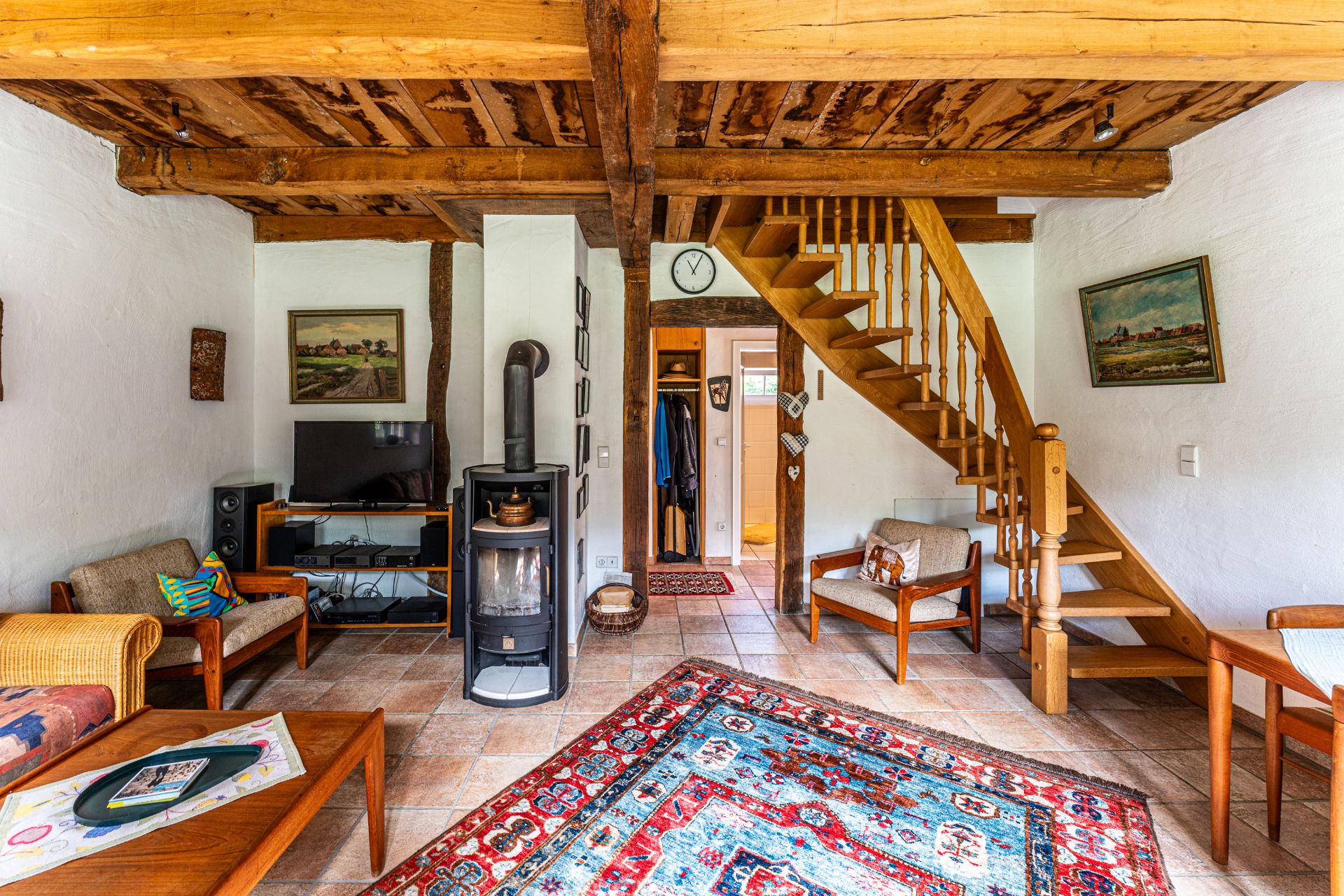 Wohnzimmer mit Holzbalkendecke, Kaminofen, Fernseher, Holztreppe und Teppich.