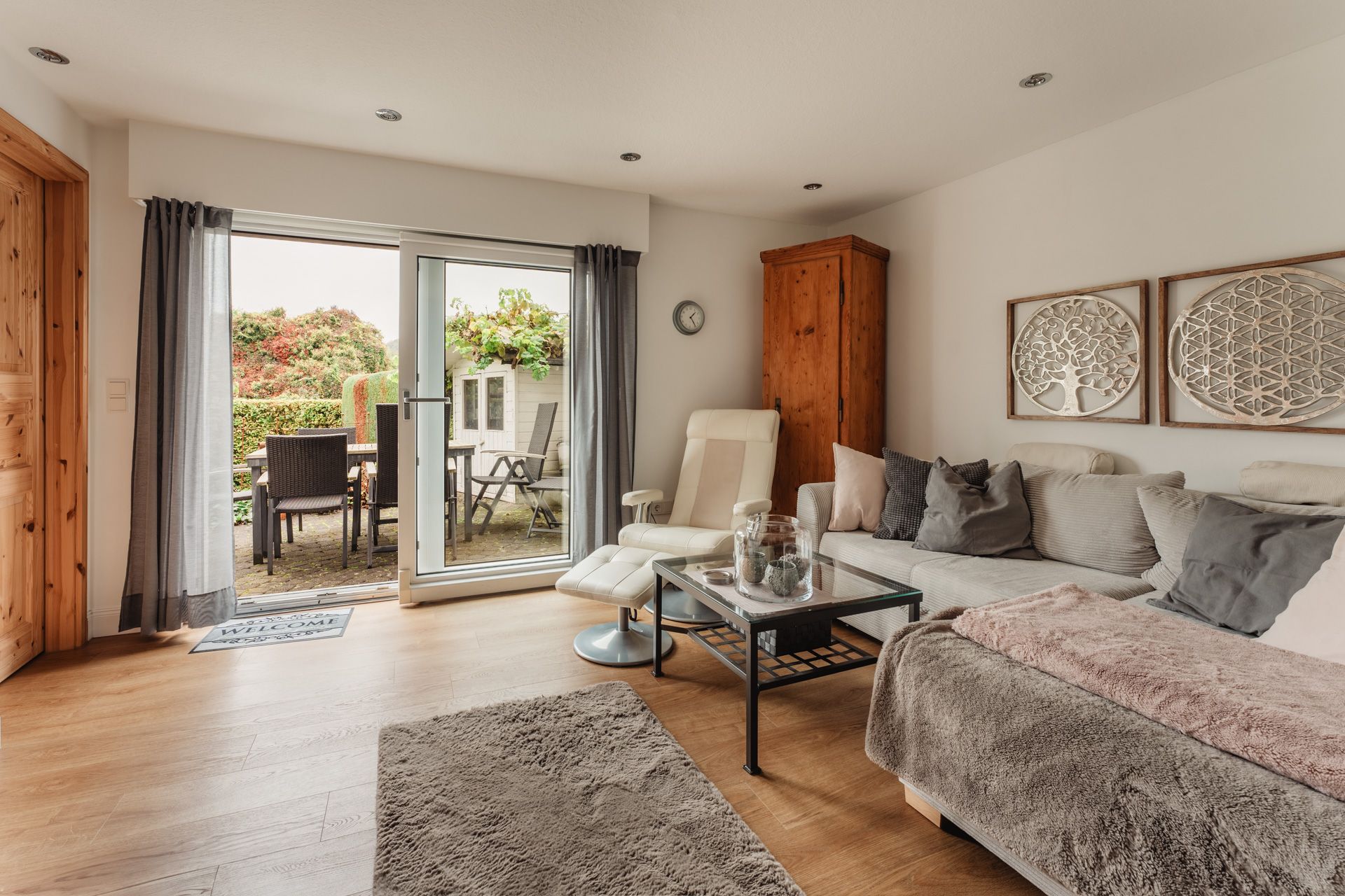 Helles Wohnzimmer mit Sofa, Sessel, Holztisch und Zugang zur Terrasse mit Gartenmöbeln.