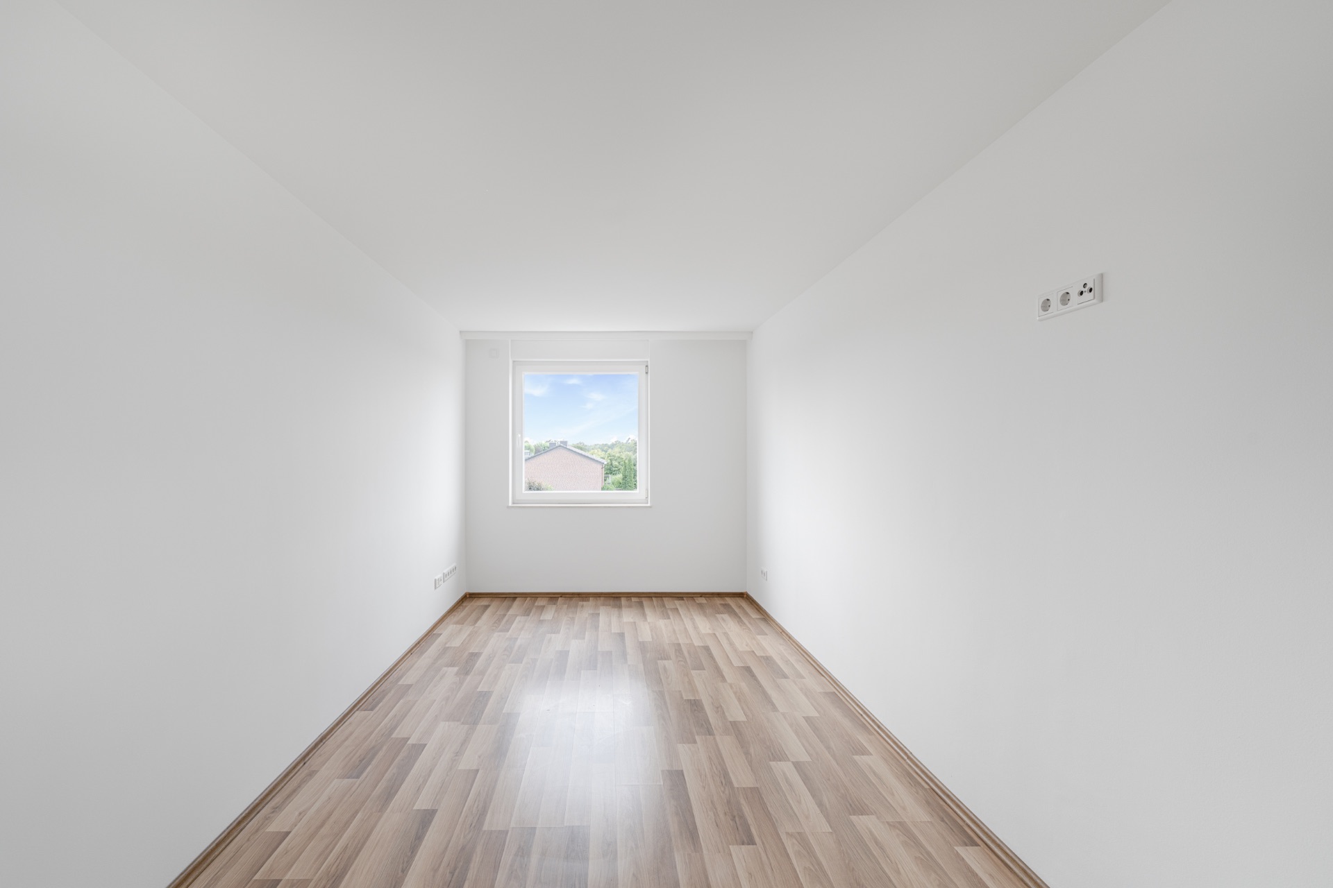 Leerer, schmaler Raum mit weiß gestrichenen Wänden, Fenster und hellem Holzfußboden.