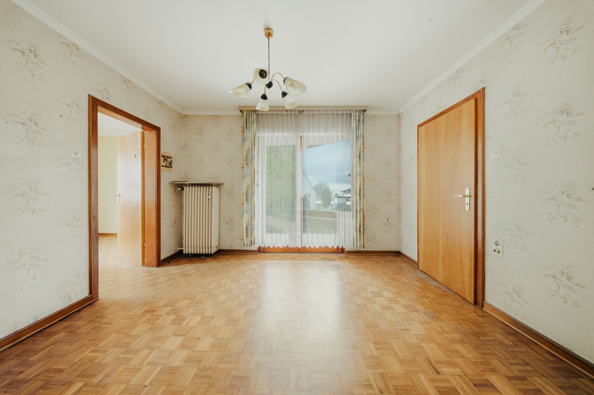 Helles Zimmer mit Parkettboden, großem Fenster, Heizung und zwei Holztüren.