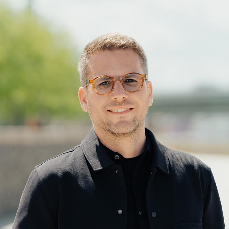 Eine Person mit Brille und dunkler Jacke steht draußen vor unscharfem Hintergrund.