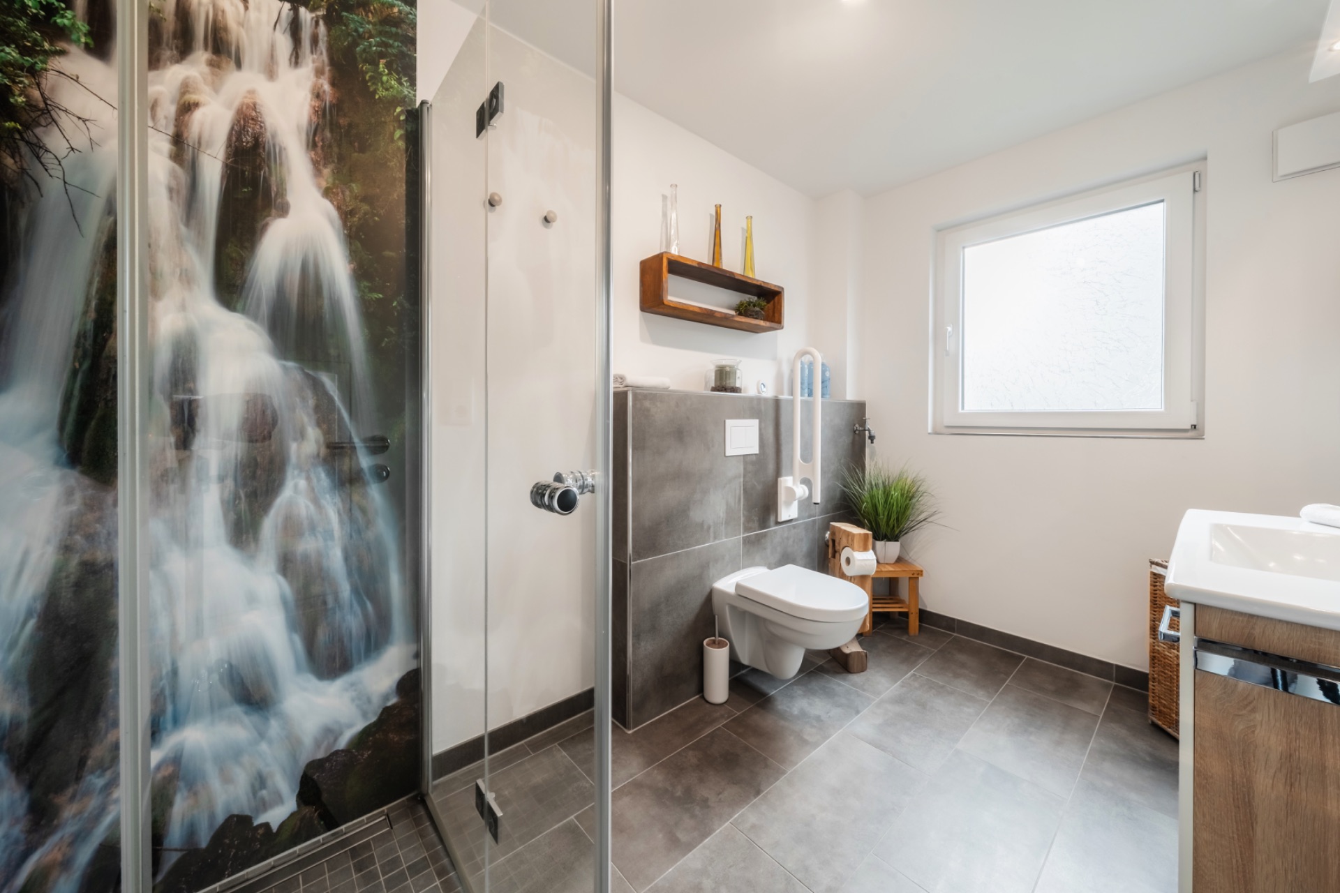 Modernes Badezimmer mit Dusche, Wasserfallmotiv, WC, Waschbecken und großem Fenster.