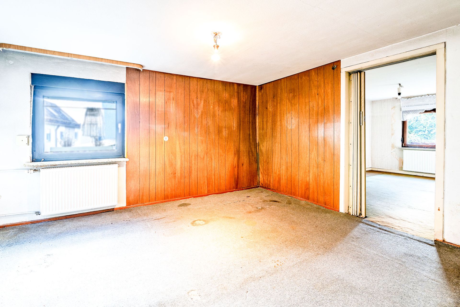 Leeres Zimmer mit Holzvertäfelung, Teppichboden, Fenster und offener Schiebetür zu angrenzendem Raum.