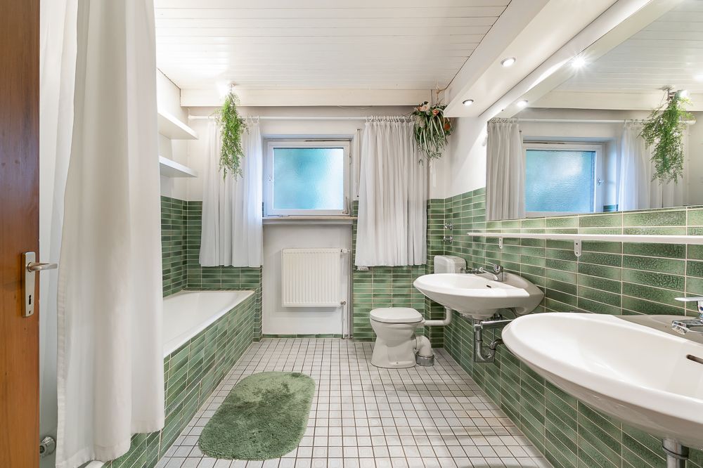 Helles Badezimmer mit grünen Fliesen, zwei Waschbecken, Badewanne und weißen Vorhängen.