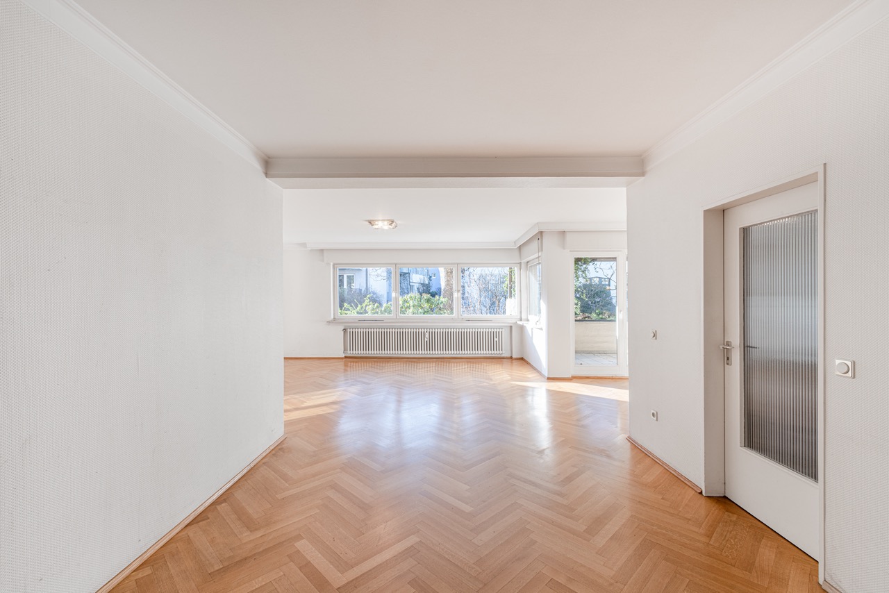 Helles, leeres Wohnzimmer mit Parkettboden, großen Fenstern und Zugang zum Balkon.