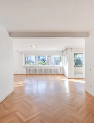 Helles, leeres Wohnzimmer mit Parkettboden, großen Fenstern und Zugang zum Balkon.