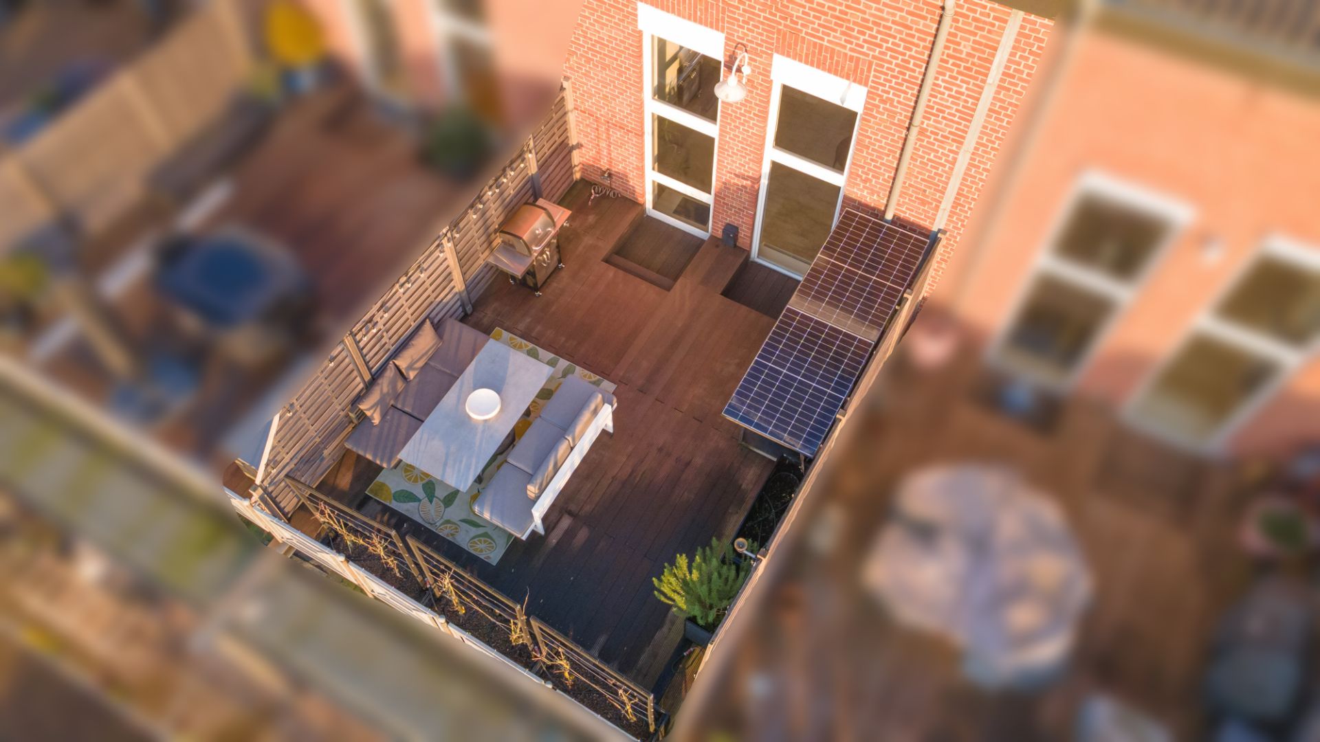 Balkon mit Holzboden, Tisch mit Stühlen, Grill und Pflanzen, angrenzend an ein Ziegelhaus.