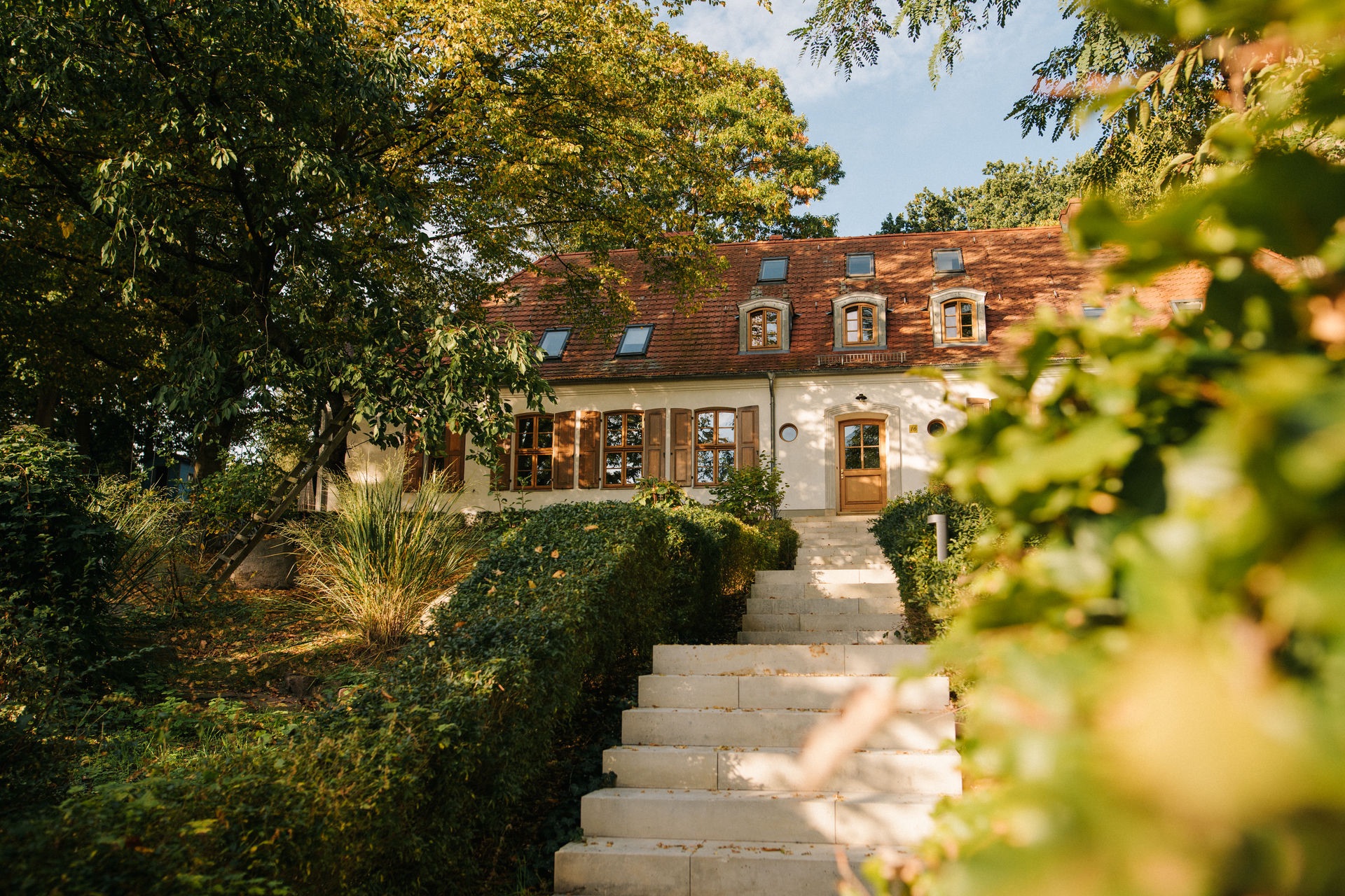 Steintreppe führt zu einem Haus mit rotem Dach und Holzfenstern, umgeben von Bäumen und Sträuchern.