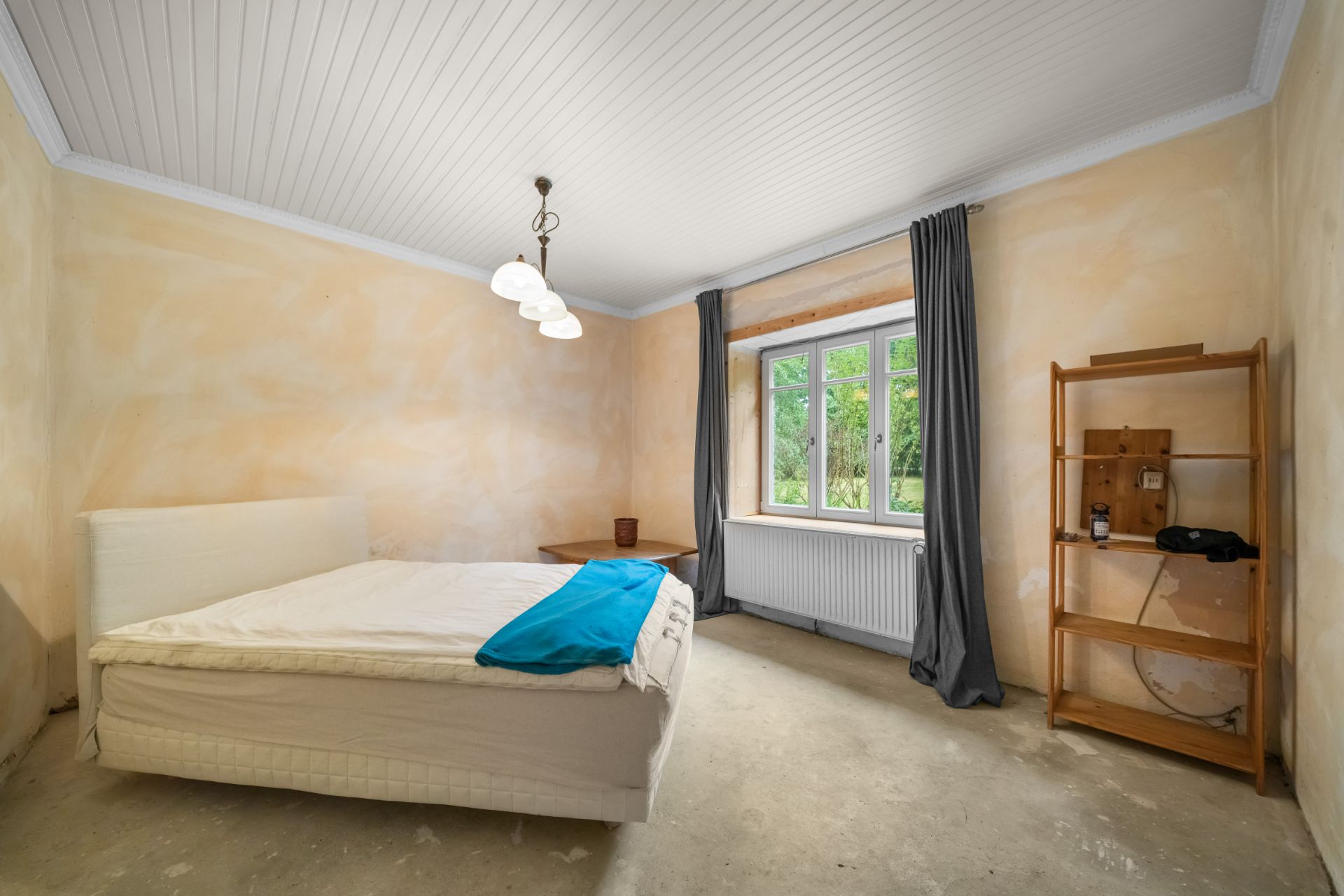 Schlafzimmer mit Bett, Regal, Tisch, Fenster mit Vorhängen und Deckenlampe.