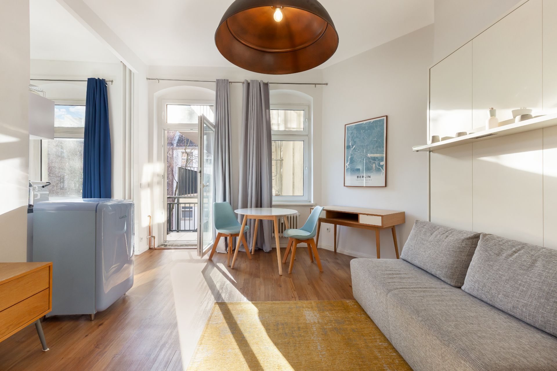 Helles Wohnzimmer mit grauem Sofa, rundem Tisch, zwei Stühlen und Zugang zum Balkon.