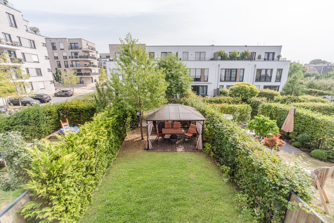 Garten mit Pavillon und Sitzgruppe, umgeben von Hecken, im Hintergrund moderne Wohngebäude.