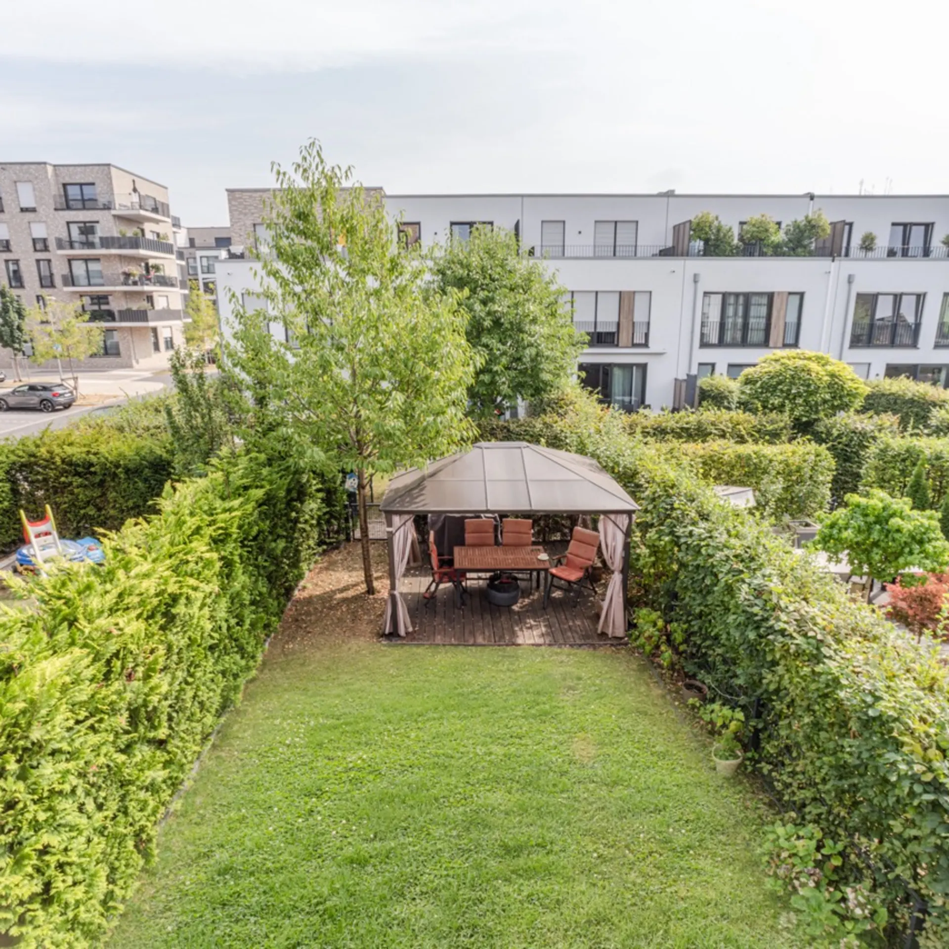 Garten mit Pavillon und Sitzgruppe, umgeben von Hecken, im Hintergrund moderne Wohngebäude.