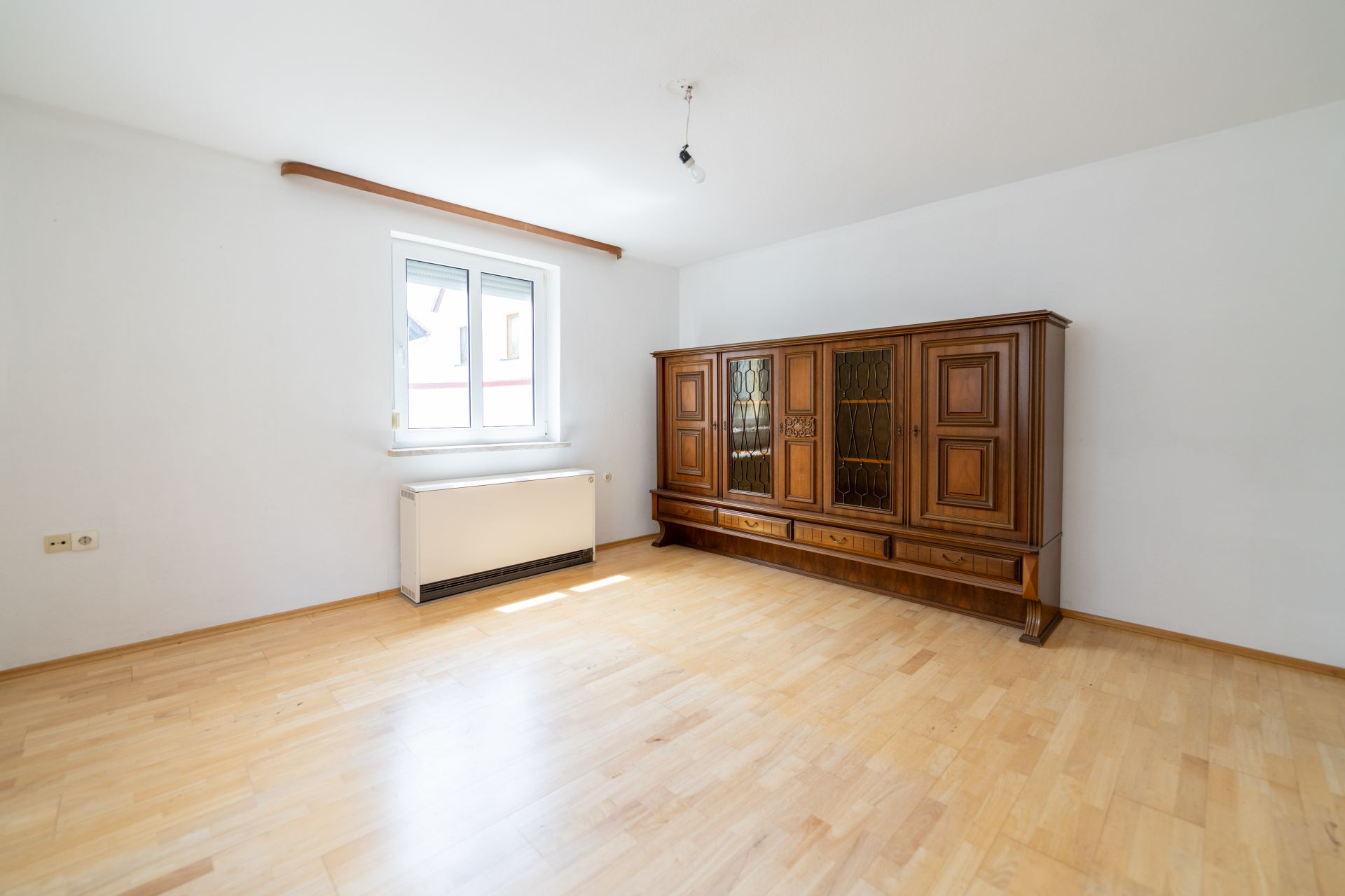 Helles Zimmer mit Holzfußboden, Fenster und großem Holzschrank an der Wand.
