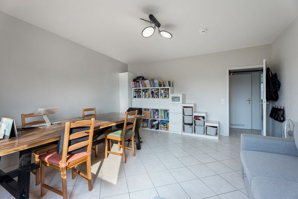 Helles Zimmer mit Esstisch, Stühlen, Regal mit Büchern und Kisten sowie grauem Sofa.