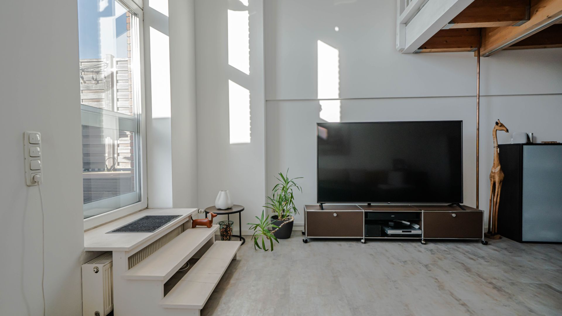 Helles Wohnzimmer mit großem Fernseher, Pflanzen und Treppe am Fenster.