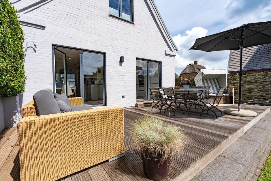 Holzterrasse mit Sofa, Esstisch, Stühlen, Sonnenschirm und Pflanze vor einem weißen Haus.