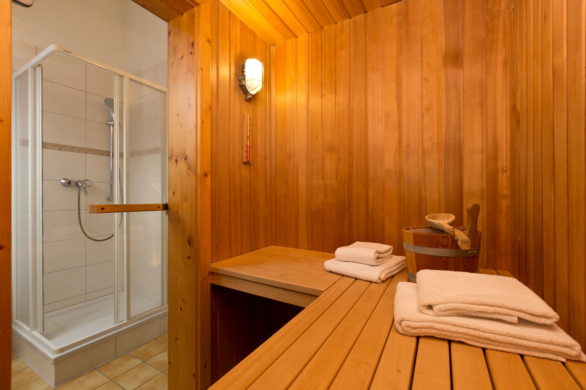 Holzsauna mit Bank, Handtüchern und einem Eimer, daneben eine Dusche mit Glastür.