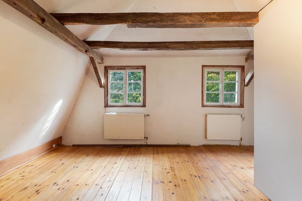 Heller Raum mit Holzboden, zwei Fenstern, sichtbaren Balken und weißen Wänden.