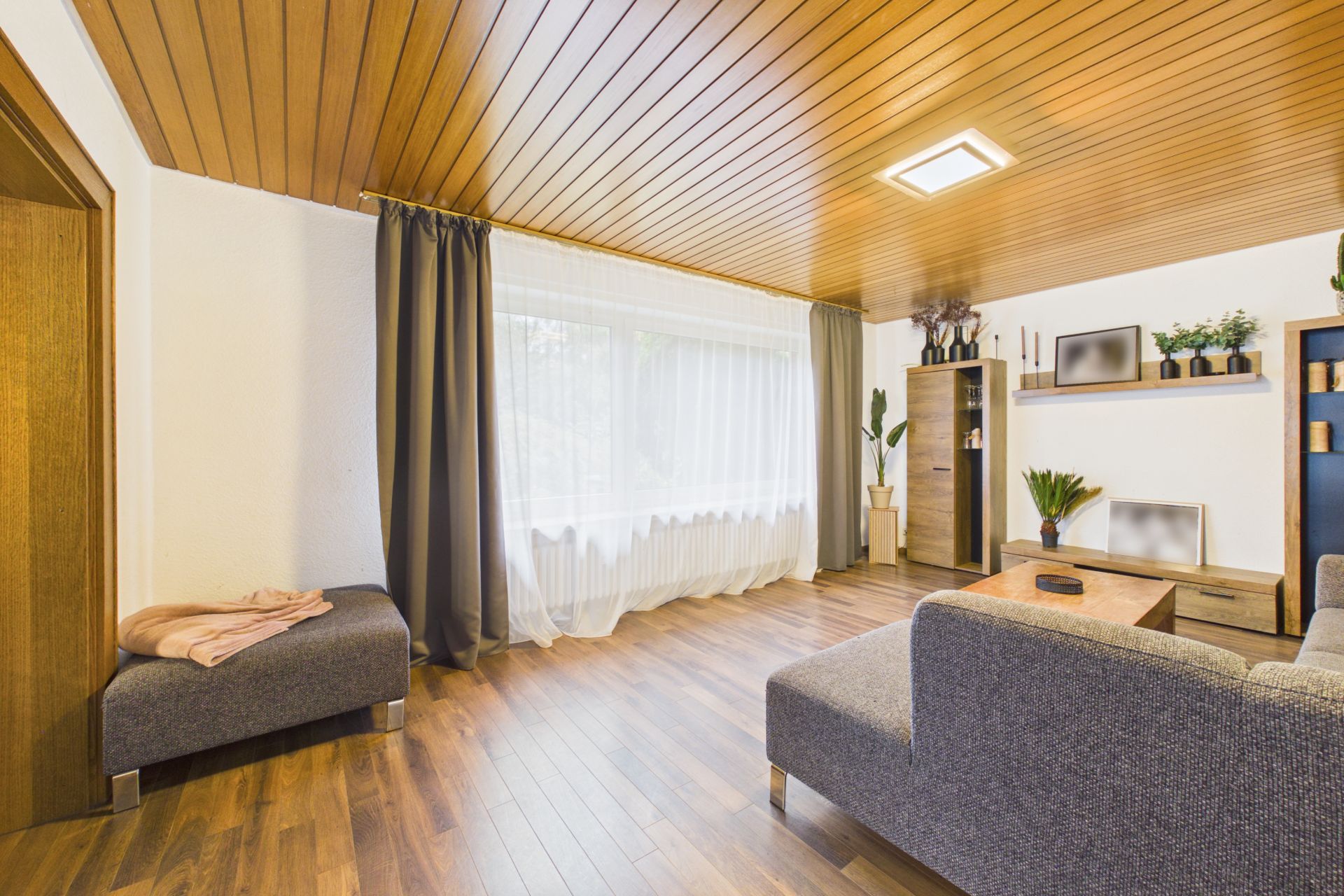 Helles Wohnzimmer mit Holzdecke, großen Fenstern, grauem Sofa und Pflanzen.