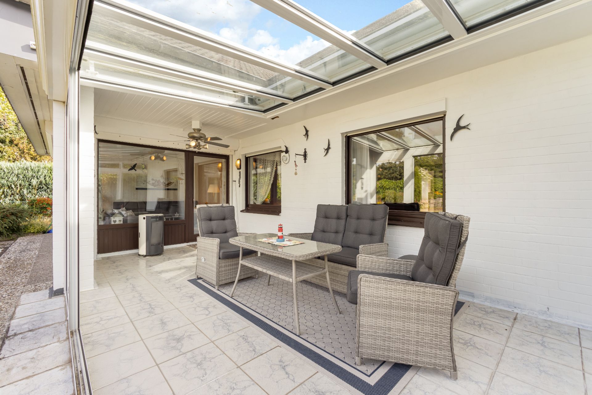 Überdachte Terrasse mit Sitzgruppe, Glastisch und dekorativen Wandvögeln an einer weißen Hauswand.