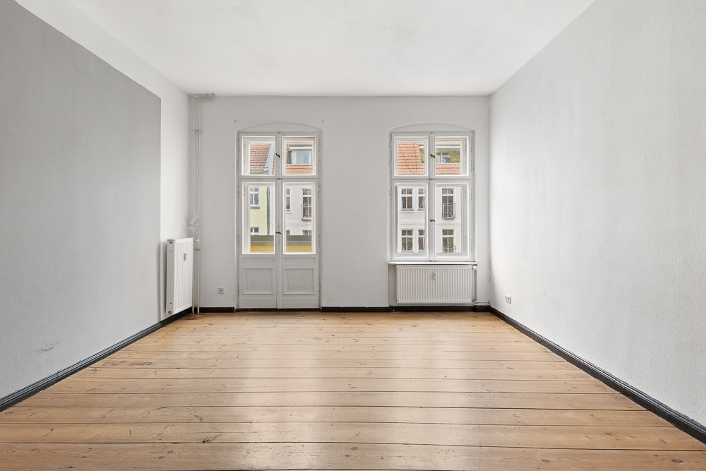 Leerer Raum mit Holzboden, weißen Wänden und zwei großen Fenstern.