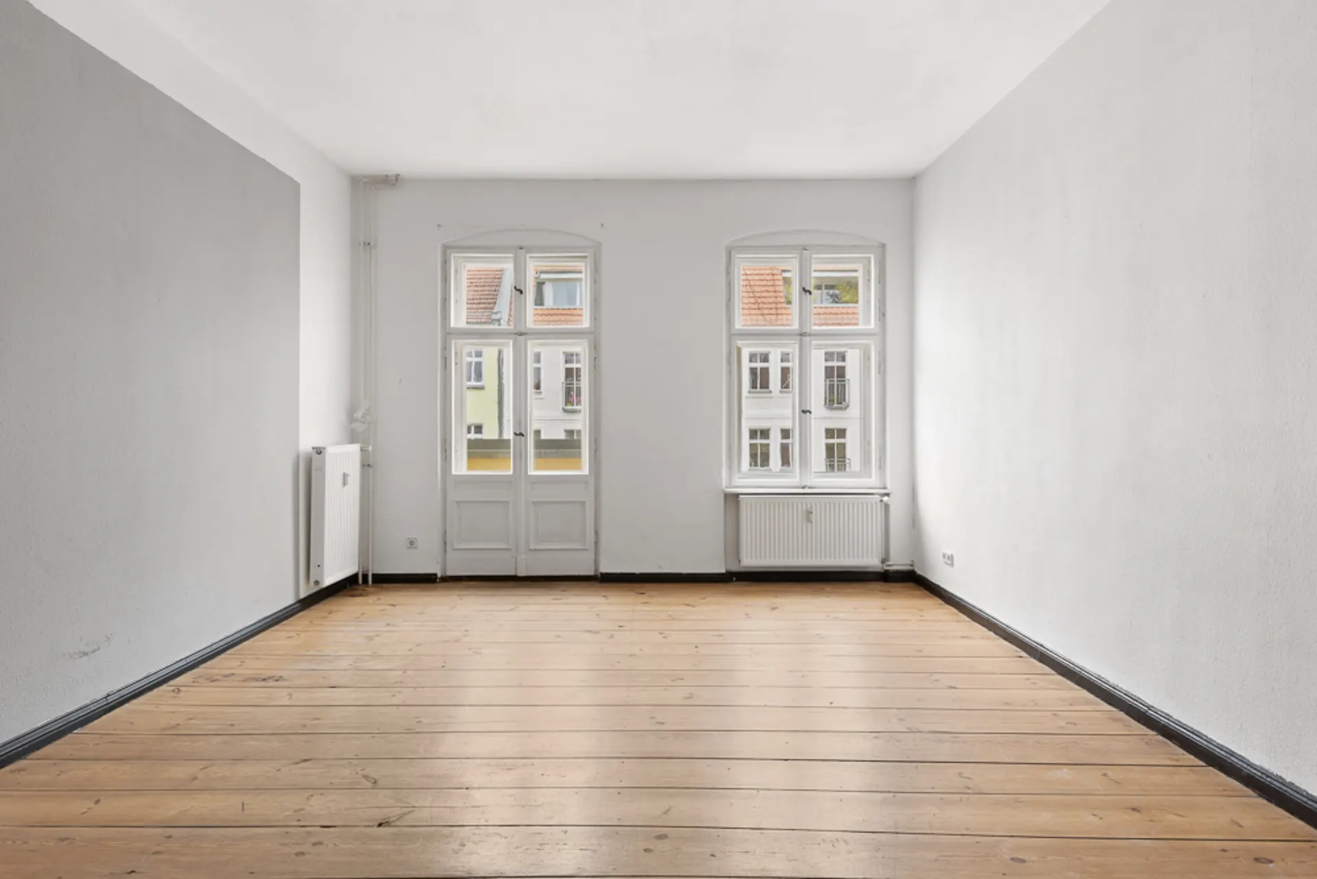 Leerer Raum mit Holzboden, weißen Wänden und zwei großen Fenstern.