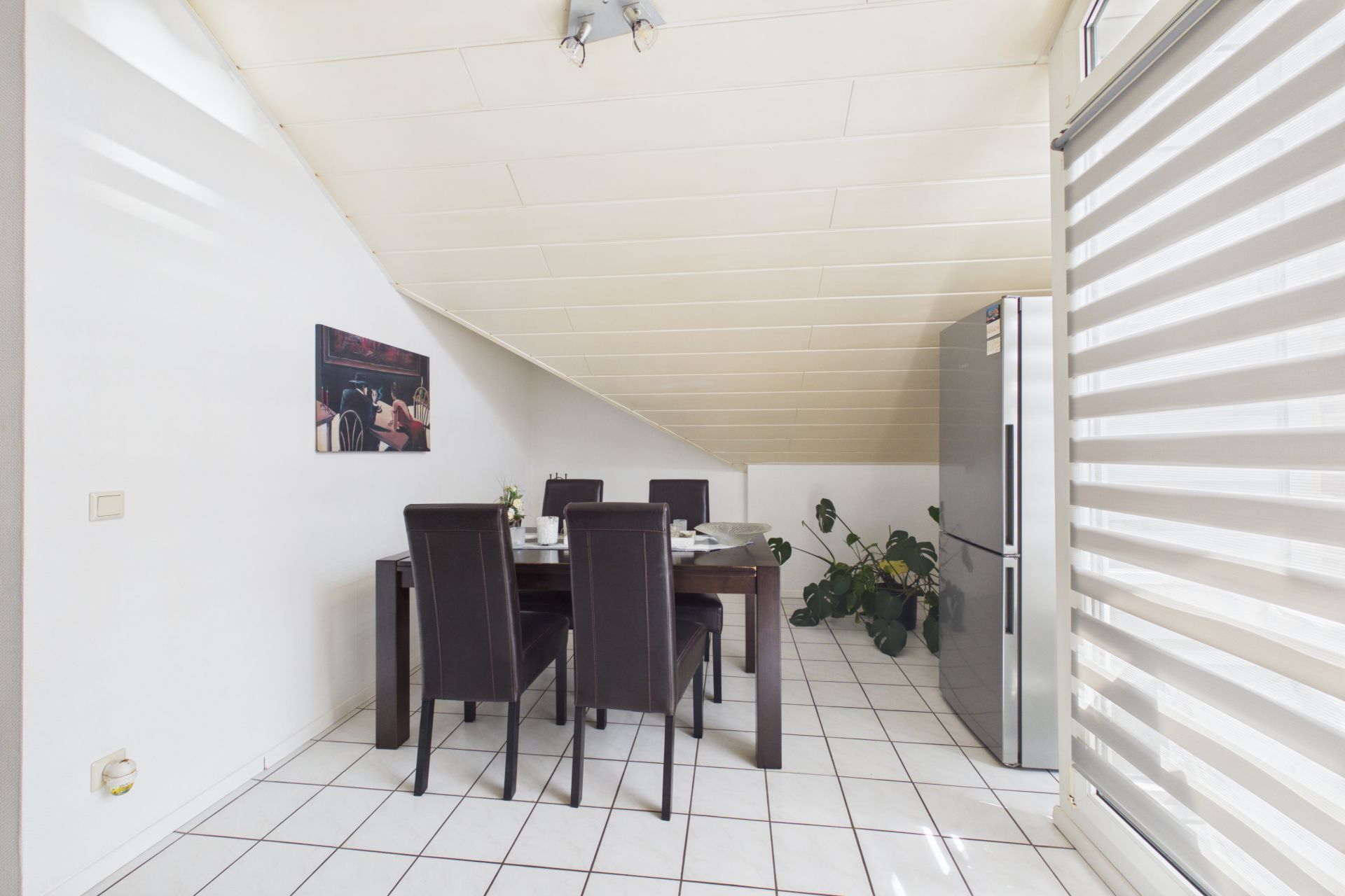 Essbereich mit dunklem Tisch, vier Stühlen, Kühlschrank und Pflanze unter schräger Decke.