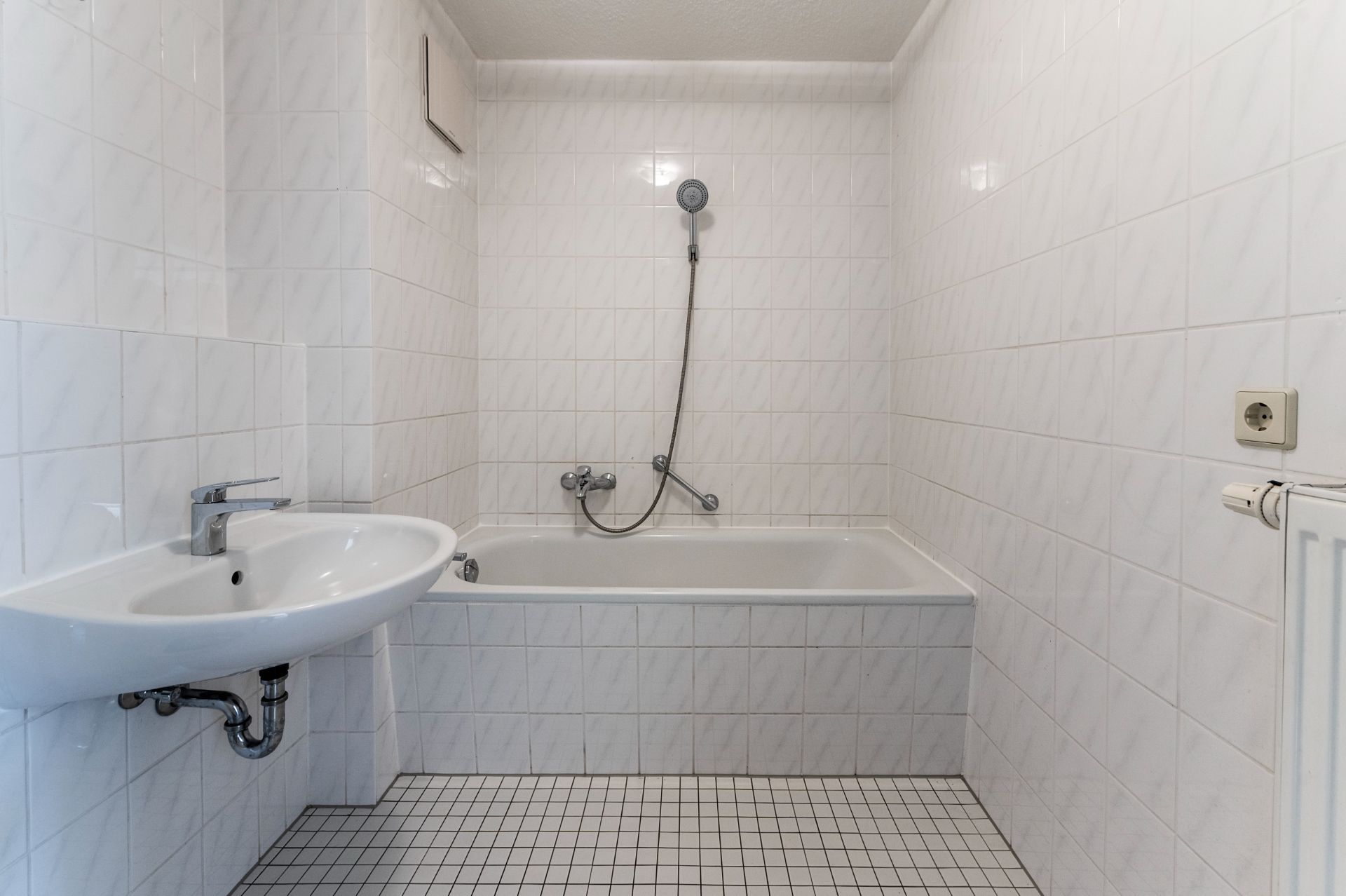 Weiß gefliestes Badezimmer mit Badewanne, Handbrause und Waschbecken.