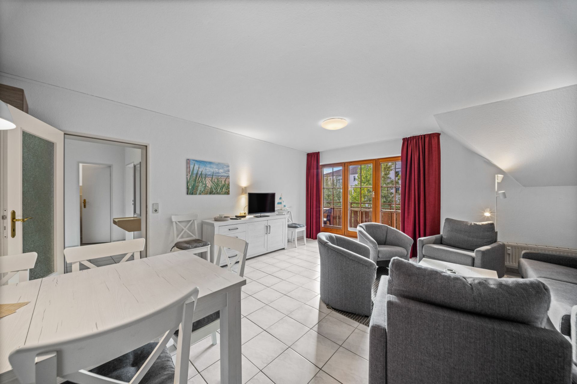 Helles Wohnzimmer mit Essbereich, grauen Sofas, TV, weißen Möbeln und Zugang zu einem Balkon mit roten Vorhängen.