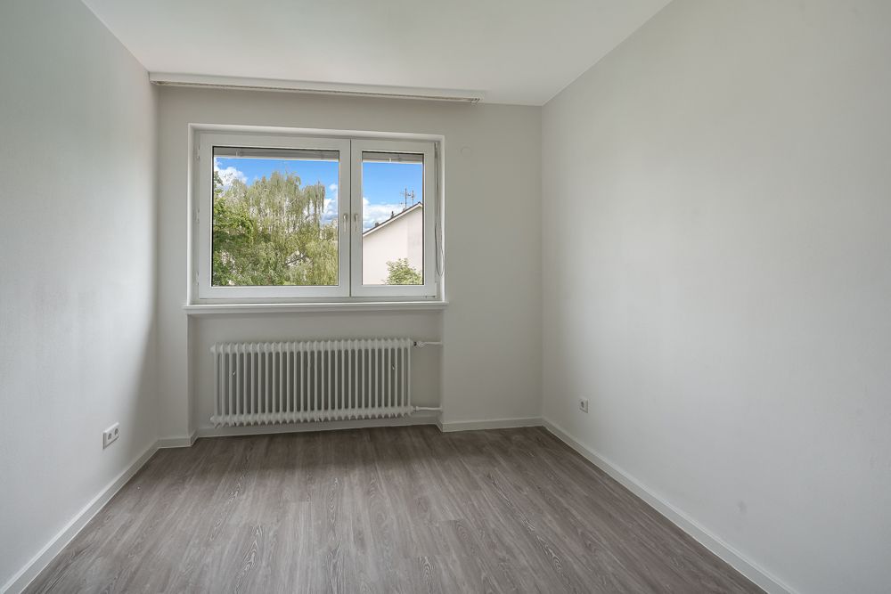 Leerer Raum mit grauem Boden, weißem Heizkörper und Fenster mit Blick auf Bäume.