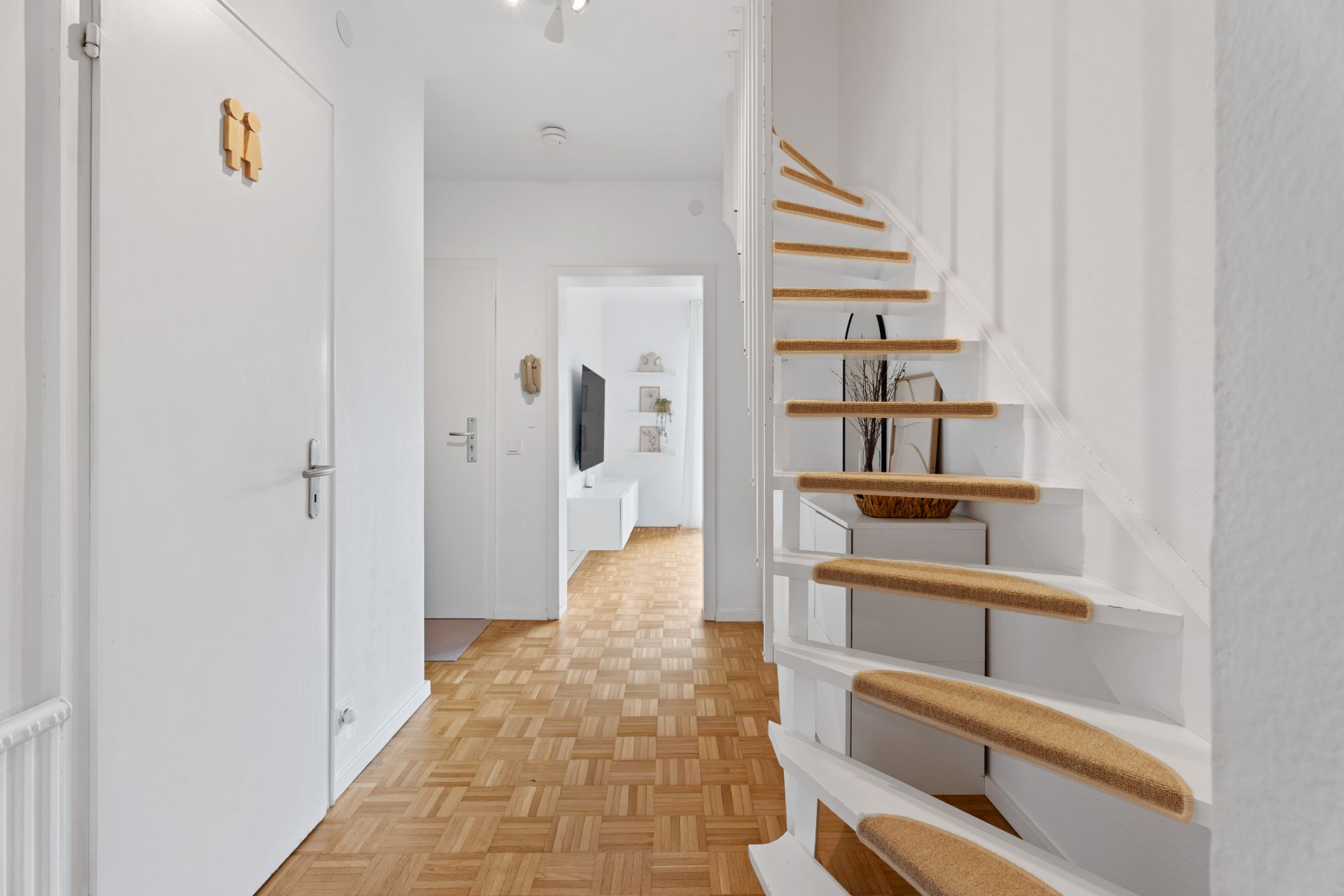 Heller Flur mit Holztreppe und Parkettboden, Blick ins Wohnzimmer mit Fernseher.