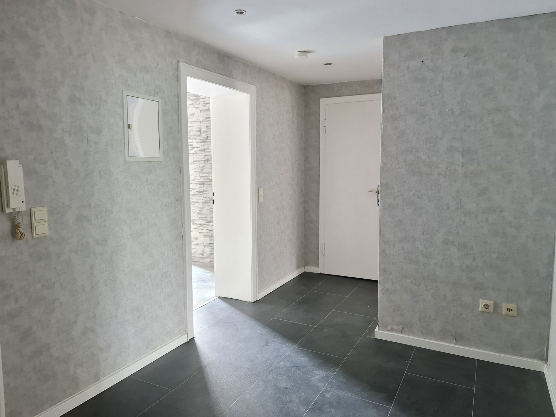 Heller Flur mit grauen Wänden, dunklem Boden, weißen Türen und Lichtschaltern.
