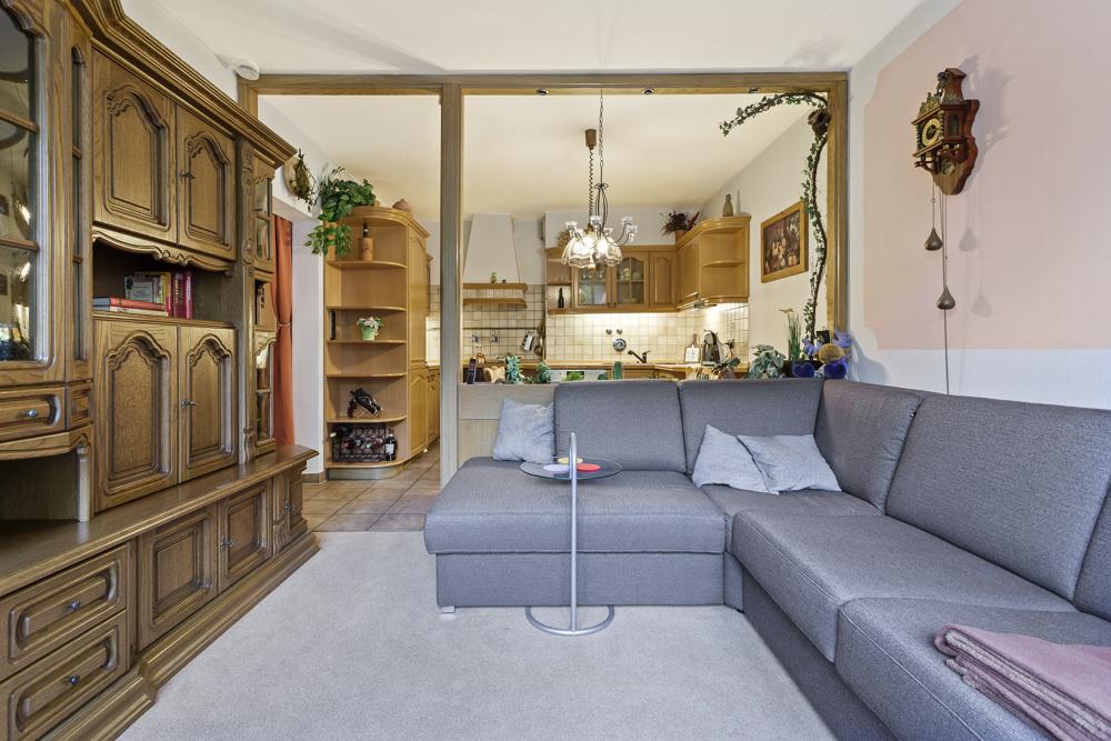 Wohnzimmer mit grauem Ecksofa, Holzmöbeln und Blick in eine offene Küche.