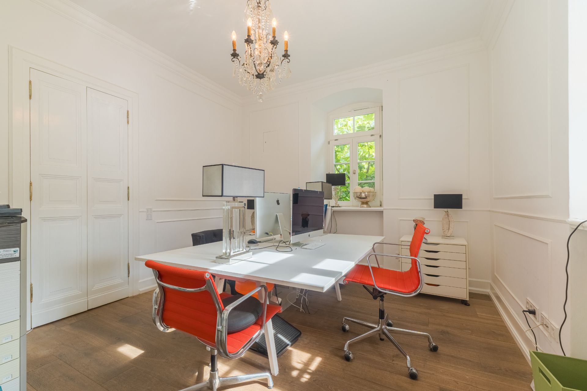 Helles Arbeitszimmer mit weißem Tisch, roten Stühlen und einem Kronleuchter unter einem Fenster.