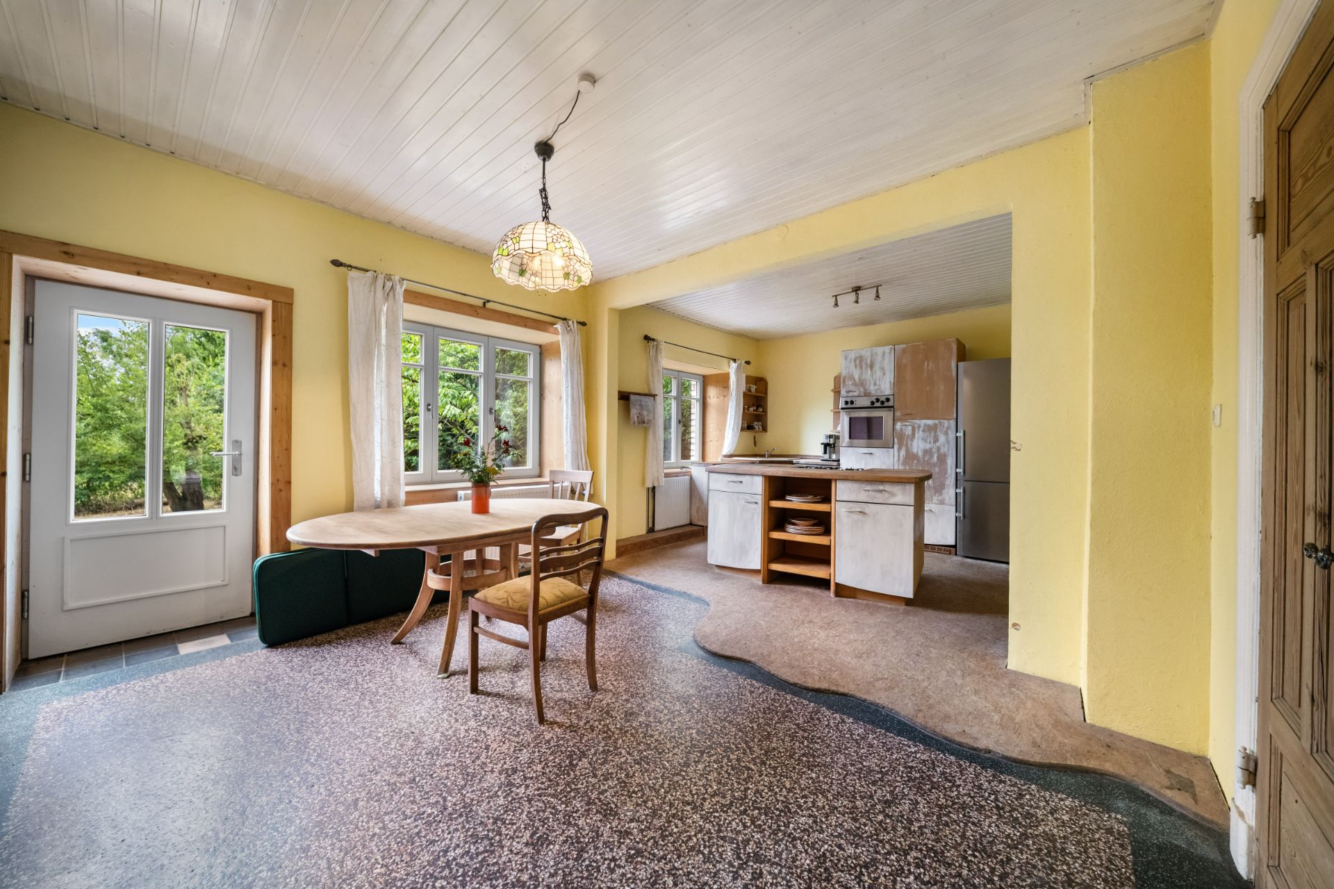 Heller Wohnraum mit Esstisch, Stühlen, offener Küche und gelben Wänden, große Fenster und Tür zum Garten.