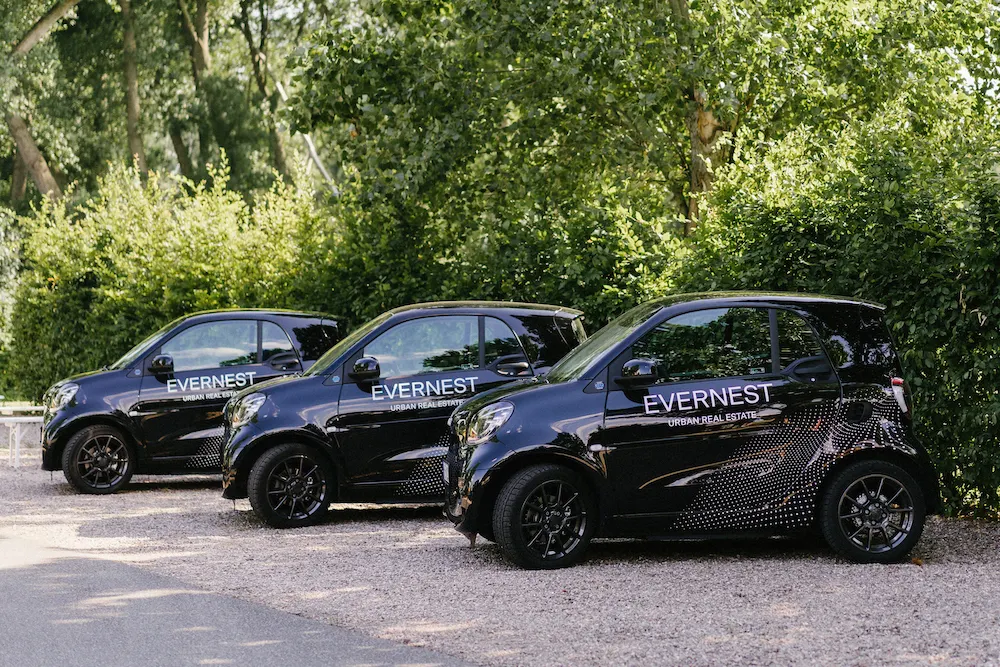 Drei schwarze Autos mit Evernest-Logo stehen nebeneinander auf einem Kiesparkplatz vor Büschen.