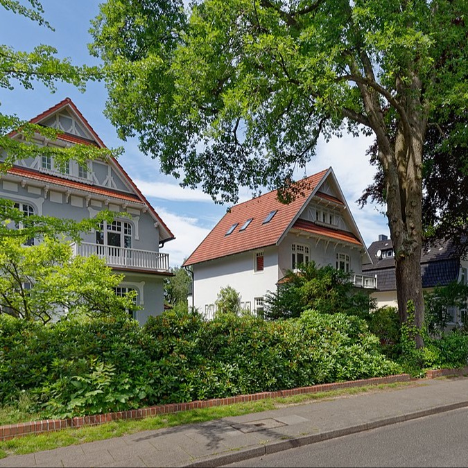 Zwei große Wohnhäuser mit roten Dächern und viel Grün an einer ruhigen Straße.