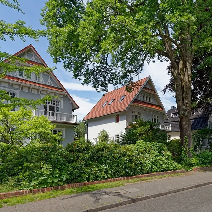 Zwei große Wohnhäuser mit roten Dächern und viel Grün an einer ruhigen Straße.