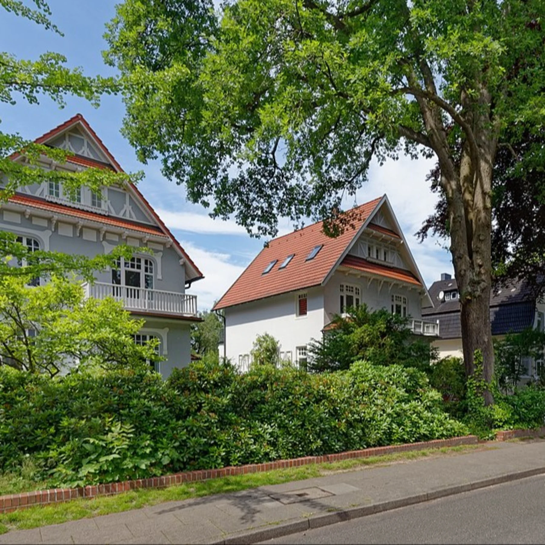 Zwei große Wohnhäuser mit roten Dächern und viel Grün an einer ruhigen Straße.