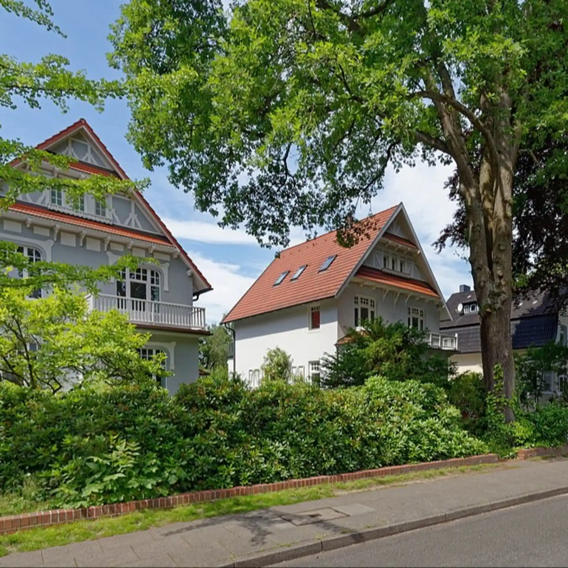 Zwei große Wohnhäuser mit roten Dächern und viel Grün an einer ruhigen Straße.