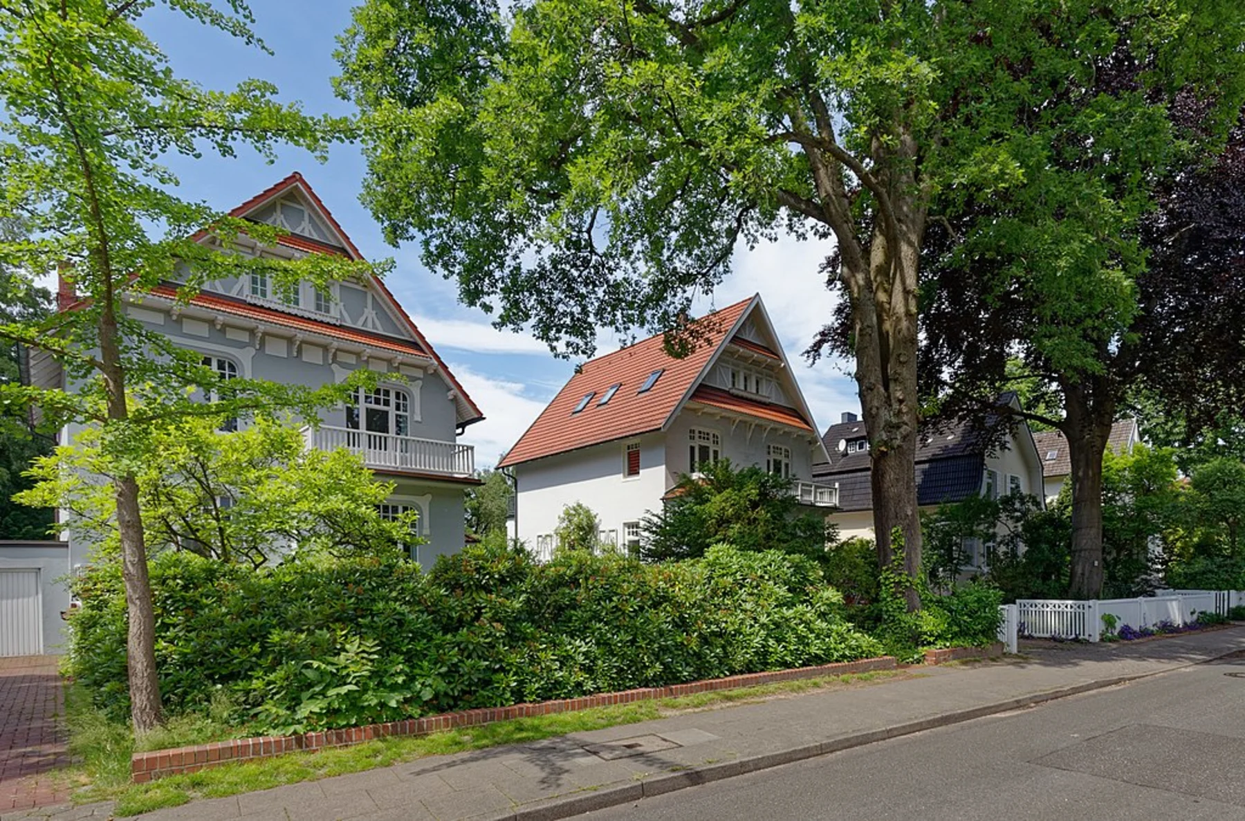 Zwei große Wohnhäuser mit roten Dächern und viel Grün an einer ruhigen Straße.